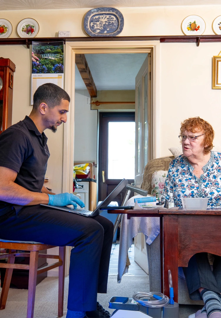 Healthcare professional consults elderly woman at home, using a laptop for patient care.