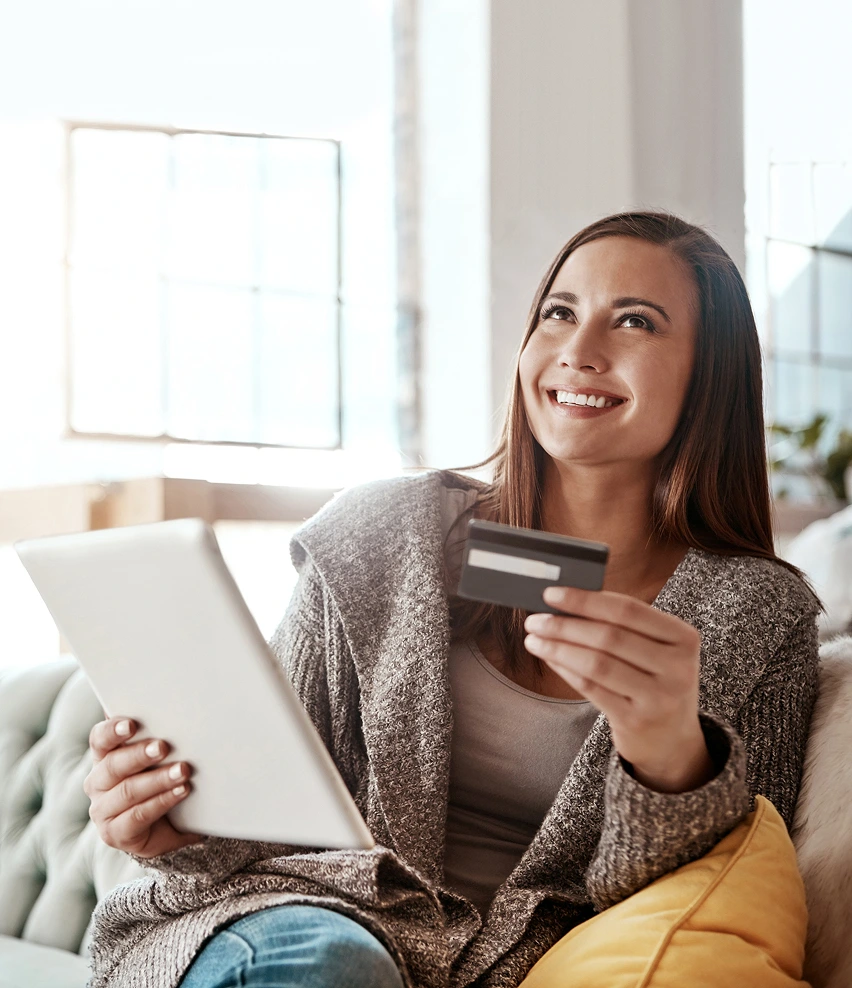 Smiling woman holding credit card and tablet, shopping online from cozy home setting.