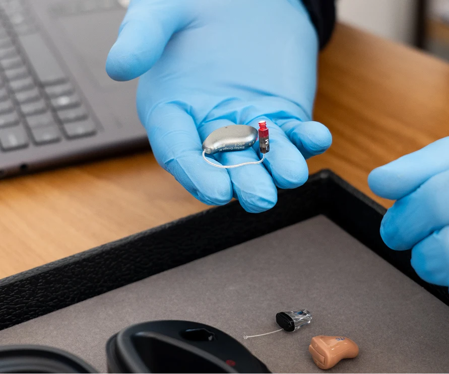 Gloved hands holding a hearing aid beside a computer, showcasing hearing support technology.