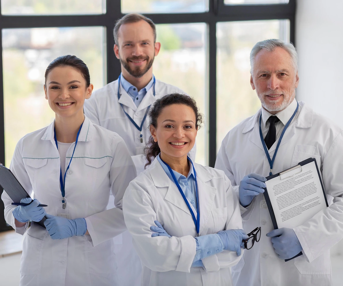 Group of four smiling healthcare professionals in lab coats holding files, standing indoors by a large window.