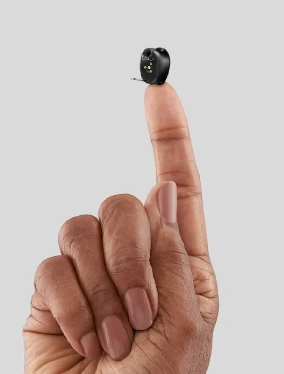 Compact black hearing aid balanced on fingertip against a neutral background.