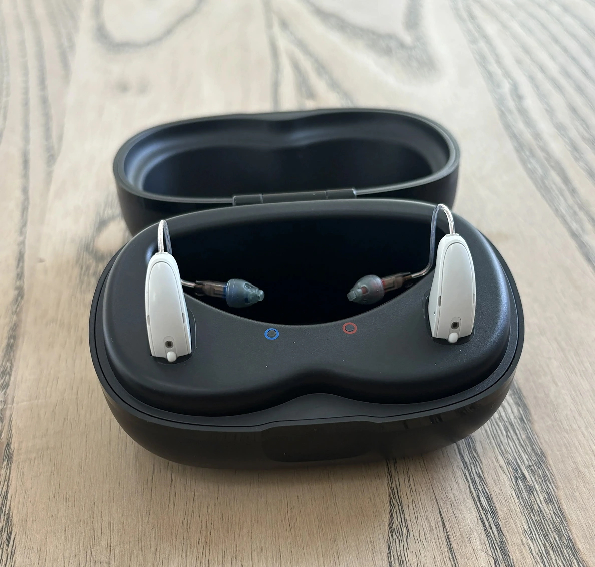 Rechargeable hearing aids in black charging case on wooden surface.