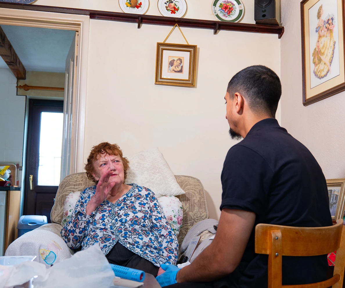 Elderly woman conversing with caregiver in a cozy living room setting, showcasing home healthcare interaction.