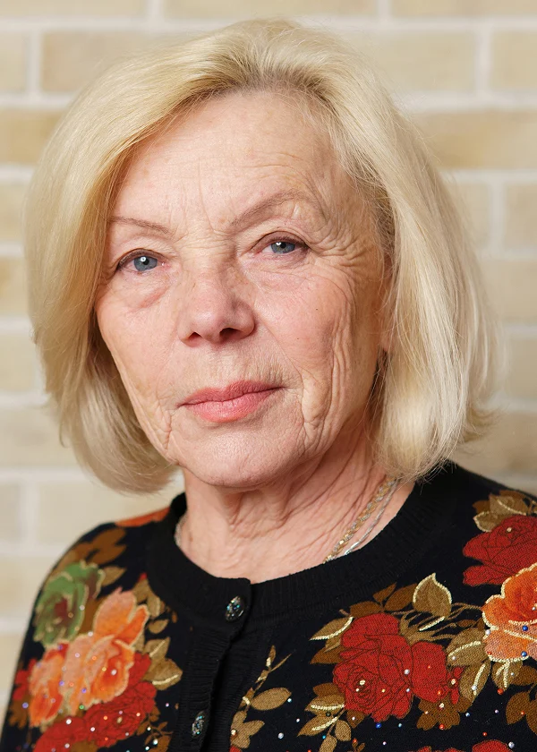Elderly woman with a blonde bob haircut in a floral sweater, standing against a brick wall background.