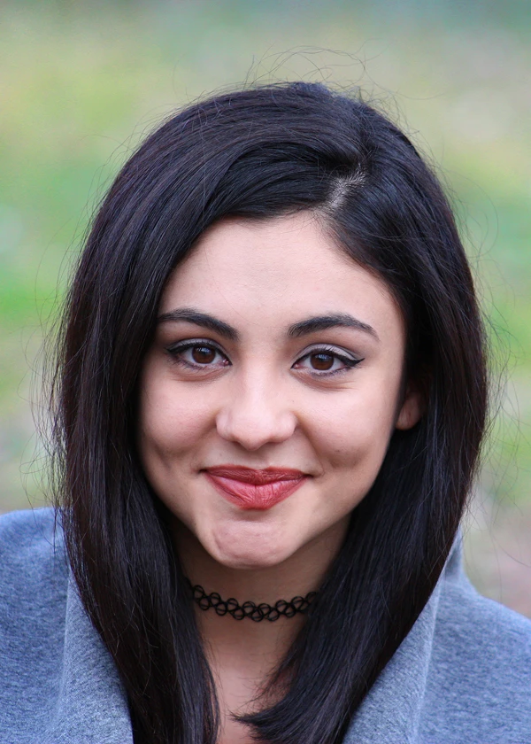 Smiling woman with dark hair and red lipstick wearing a gray coat outdoors.