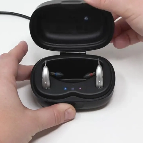 Hearing aids in a black charging case held by hands on a white background.