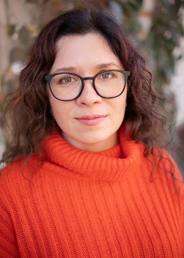 Woman wearing glasses and orange sweater, standing outdoors with soft background blur.