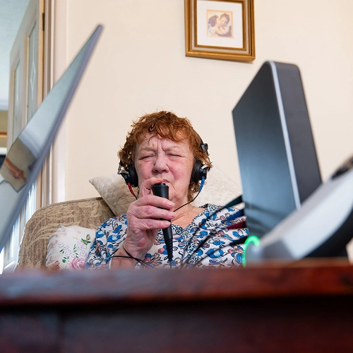 Elderly woman singing with headphones and microphone in home setting, surrounded by technology.