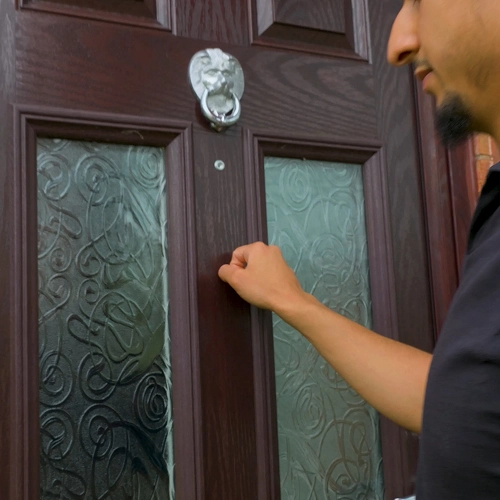 Person knocking on a wooden front door with decorative glass panels and a silver knocker.