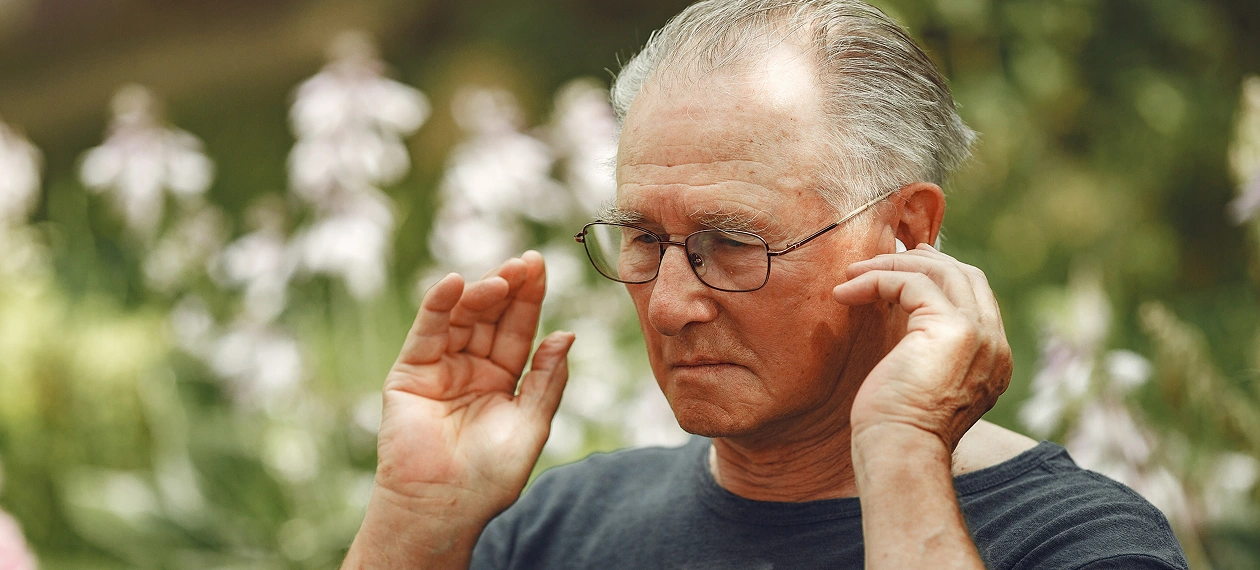 Elderly man practicing relaxation in garden, focusing and holding hands near head, surrounded by blurred flowers.