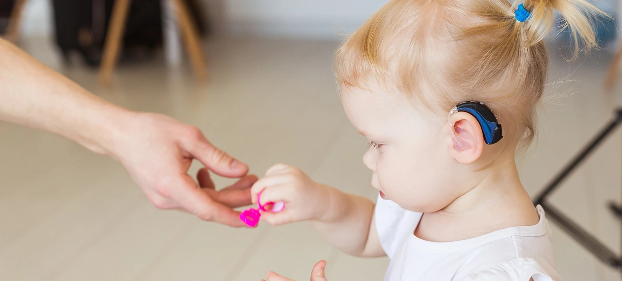 Toddler with a hearing aid holding a pink toy, interacting with a person's hand in a bright room.