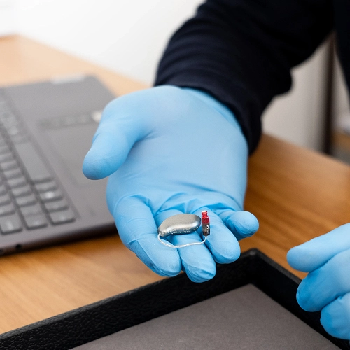 Person with blue gloves holding a hearing aid device near a laptop on a wooden desk.