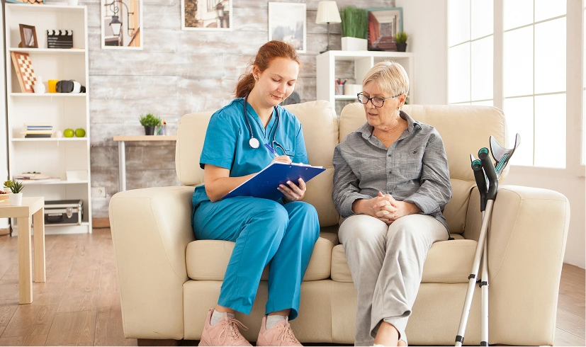 Nurse discussing medical care with senior woman on a sofa, with crutches nearby, in a bright living room setting.