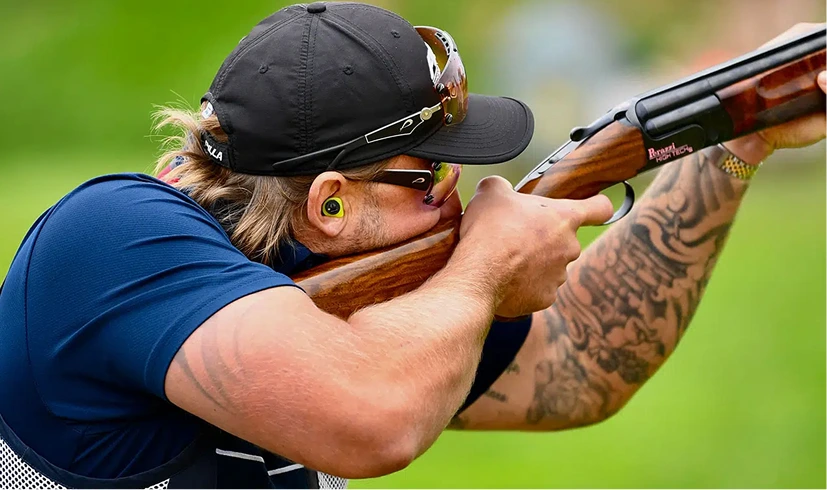 Man aiming shotgun during shooting competition, wearing protective glasses and earplugs, with tattoos visible on arms.