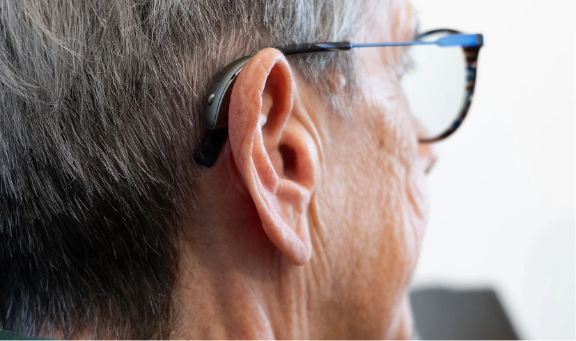 Close-up of a person with gray hair wearing a hearing aid behind the ear and eyeglasses.