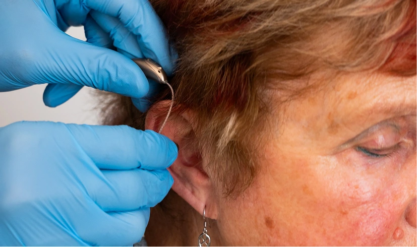 Fitting a hearing aid on an elderly woman's ear, showing careful adjustment by hands in gloves.