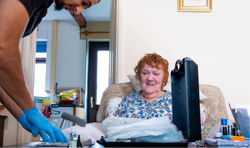 Caregiver visiting smiling elderly woman at home, preparing medical supplies on table.
