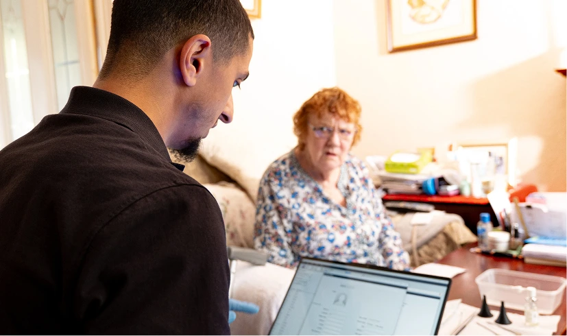 Healthcare worker using laptop while consulting elderly woman at home.