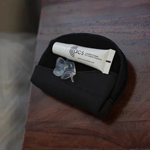 Earplugs and cream on wooden surface, with a black pouch; ACS Custom Comfort accessories for hearing protection.
