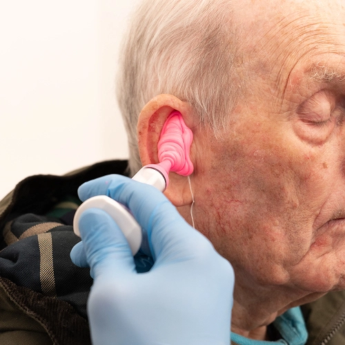 An audiologist creates an ear mold for a hearing aid in an elderly man's ear using a pink substance.