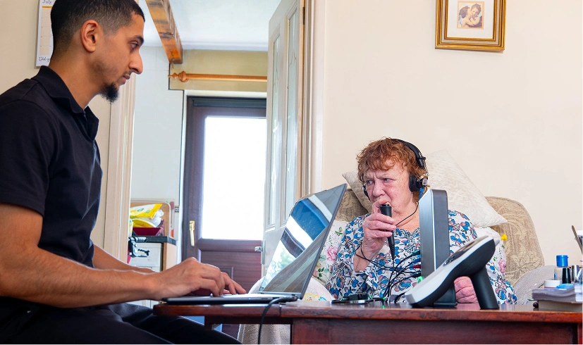 Young man assists elderly woman with telehealth call, using a laptop and headset at home.