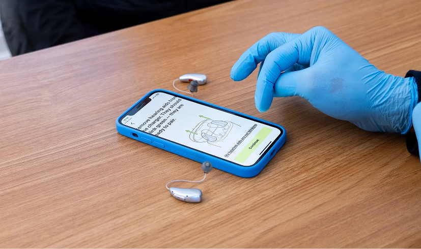 Gloved hand operates smartphone displaying hearing aid app, with two hearing aids on wooden table.