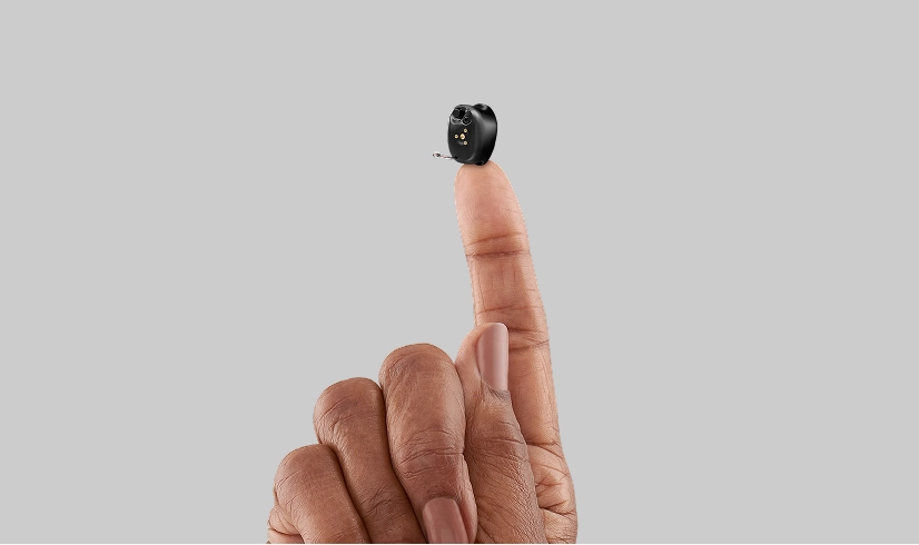 Hand holding a small black hearing aid on fingertip against a gray background, highlighting its compact design.