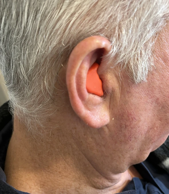 Elderly man with hearing protection earplug inserted in left ear for noise reduction and safety.