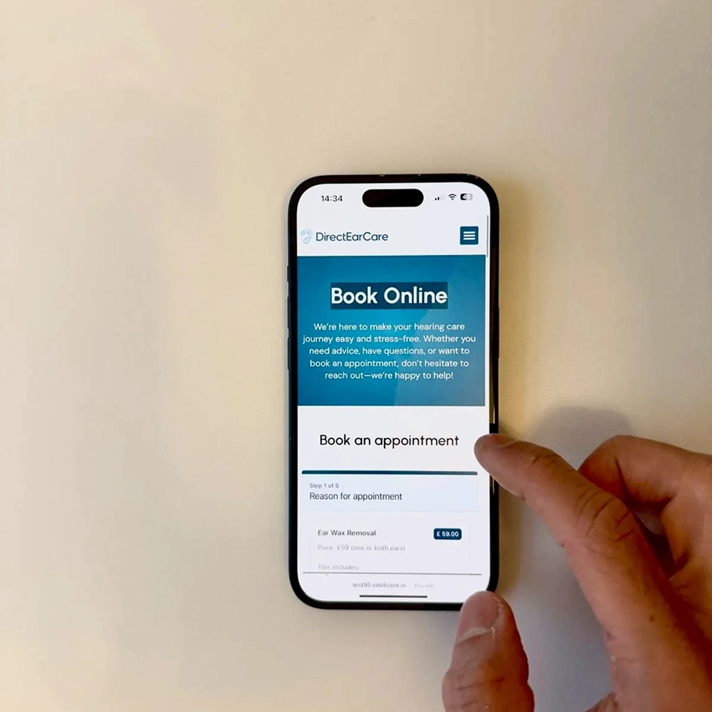 Person using a smartphone to book an online hearing care appointment with Direct Ear Care website visible.