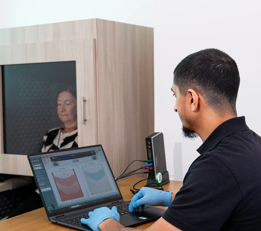 Technician conducting audiometry test in soundproof booth with patient, using specialized software on laptop.