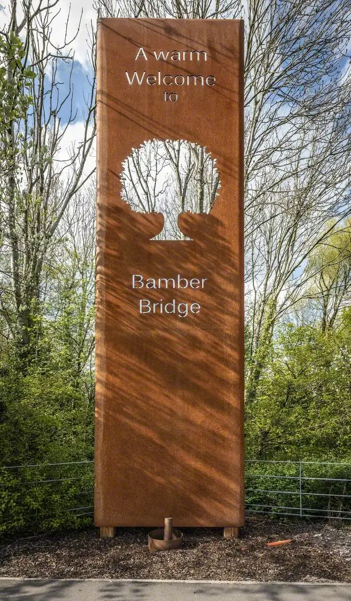 Sign saying A warm welcome to Bamber Bridge with a tree cutout, set against a backdrop of trees.