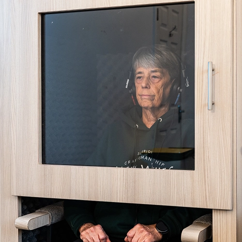 Elderly woman undergoing hearing test in soundproof booth, wearing headphones, focused expression.