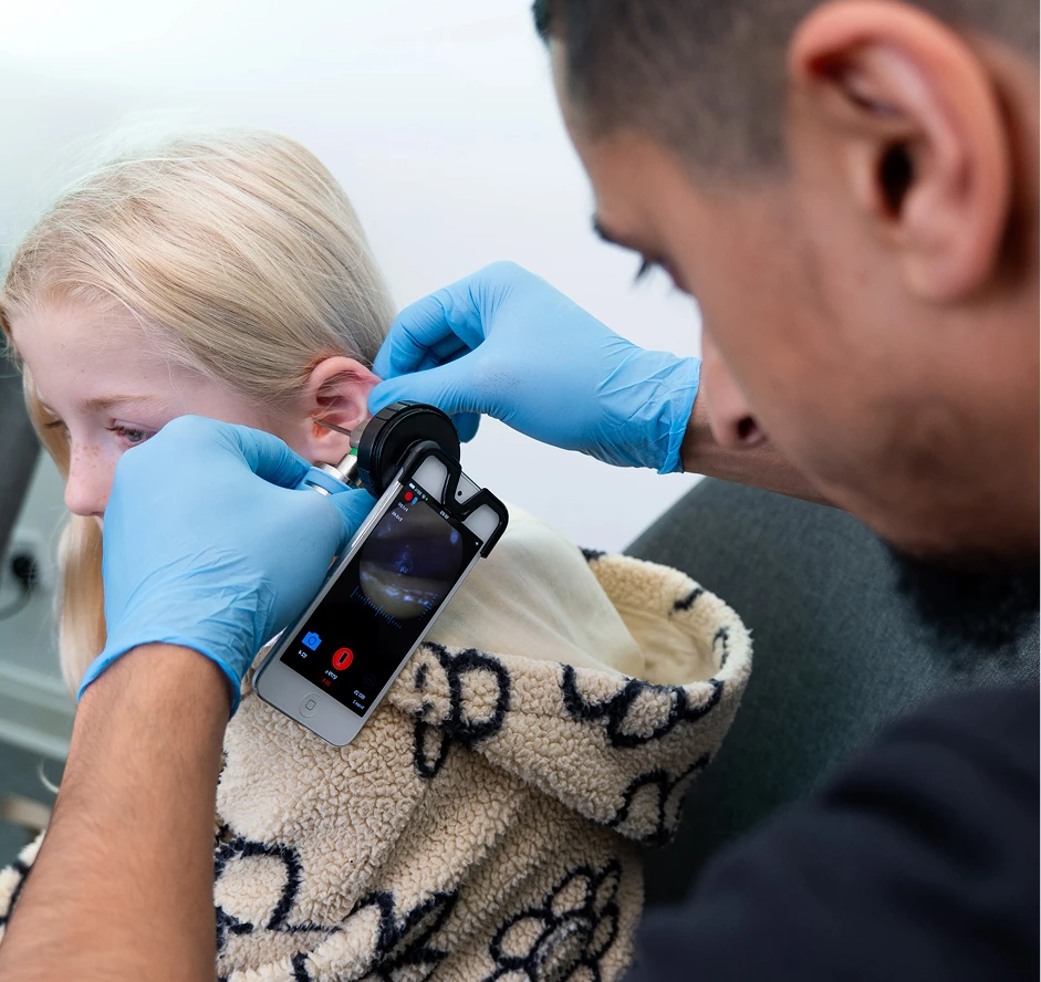 Doctor examines child's ear with smartphone otoscope, using blue gloves.