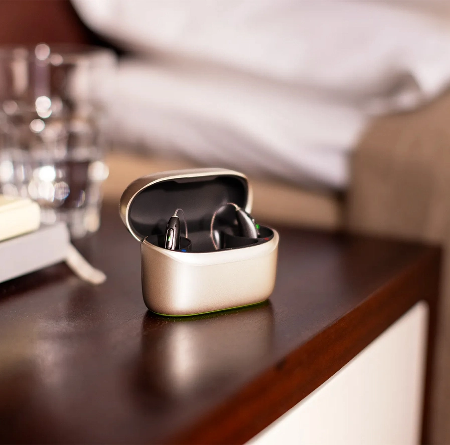Sleek wireless earbuds with charging case on a wooden table beside a bed in a cozy bedroom setting.