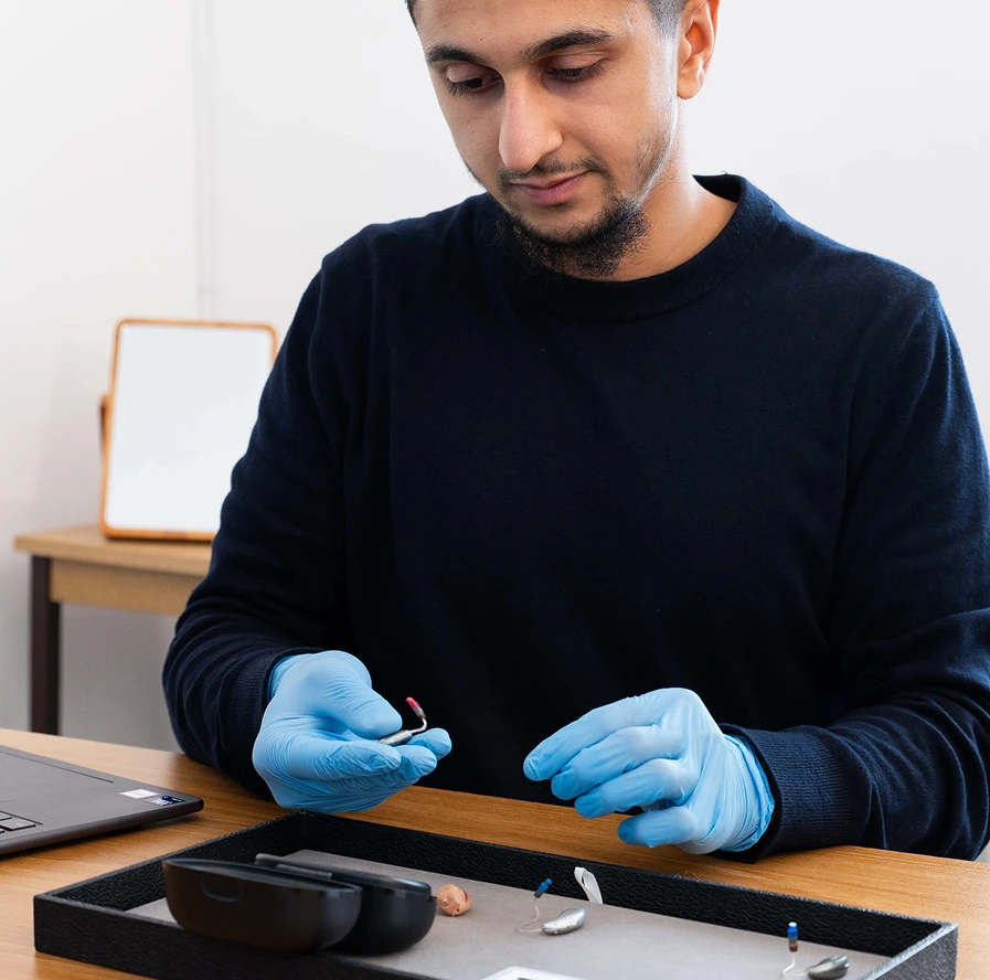 Technician wearing gloves assembles a hearing aid at a desk with tools and a laptop.