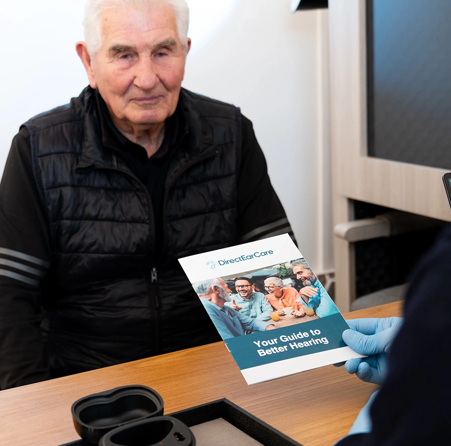 Elderly man receiving a hearing guide pamphlet at a hearing care consultation desk.