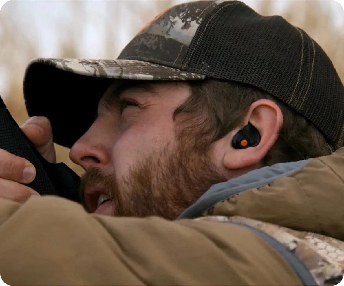 Man wearing camo hat takes aim using binoculars, sporting wireless earbuds for outdoor adventure.