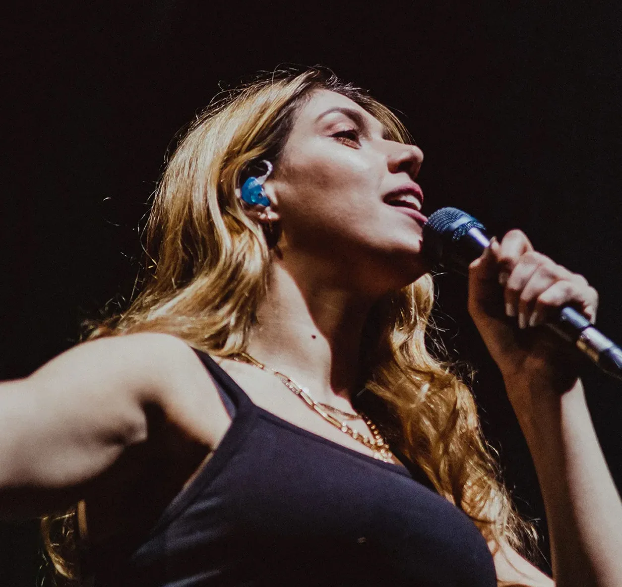 Singer with long hair performing live on stage holding a microphone, wearing a black tank top.